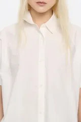 Camisa blanca de corte oversize confeccionada en 100% algodón, con cuello camisero, manga corta recta y ancha, cierre frontal con botones y pespuntes a contraste.