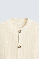 Chaleco color crema de punto con estructura, cuello redondo, sin mangas y cierre frontal con botones marrones.
