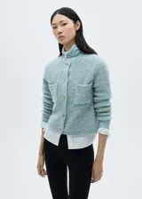 Chaqueta color celeste de tejido bouclé con cuello redondo, cierre frontal con botones efecto joya, dos bolsillos de parche frontales y mangas largas.