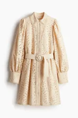 Vestido corto color beige con broderie, cuello camisero, botonadura frontal, cinturón extraíble con hebilla redonda, mangas globo largas con puños abotonados y ribetes festoneados.