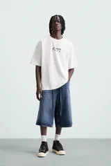Camiseta blanca de corte holgado, cuello redondo y manga corta. Estampado frontal y trasero con gráficos y textos combinados en color negro.
