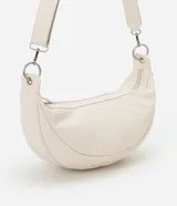 Bolso mediano color crema, con forma de media luna y correa ajustable con aros metálicos.