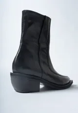 Bota corta negra de cuero con caña ajustada, punta cuadrada y taco ancho y bajo. Presenta un cierre lateral y costuras visibles en el empeine.