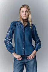 Camisa de calce recto confeccionada en tencel azul denim, con manga larga amplia con frunces en los hombros y cuello camisero. Presenta bordado de hilo en mangas y cartera. Mangas terminadas en puño con botón y ojal. Cartera delantera con botones metálicos con el logo de la marca.