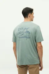 Remera verde de manga corta con cuello redondo y estampado en la espalda con la frase "Côte d'Azur Surfer avec les mouettes" y un dibujo de una sombrilla, una reposera y gaviotas.