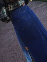 Falda larga de jean azul oscuro, corte amplio y recto, con abertura frontal y terminación desflecada en el ruedo. Presenta un pequeño parche de cuero marrón en la abertura lateral.