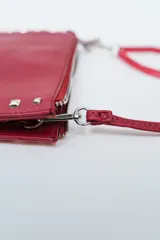 Cartera pequeña tipo clutch o bandolera de color negro, con borde decorado con tachas cuadradas plateadas. Posee cierre superior con cremallera y correa larga ajustable para llevar al hombro o cruzada.