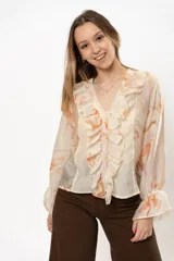 Camisa color crema con estampado floral en tonos naranja, con cuello en V y volados frontales. Mangas largas abullonadas con puños elásticos.