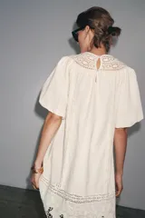 Vestido corto color crudo confeccionado en hilatura con mezcla de algodón y lino. Presenta cuello redondo, manga corta abullonada, detalle de bordados a tono y bajo recto. Cierre en espalda con abertura y botón.