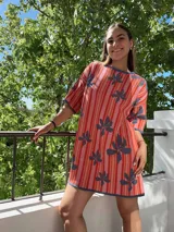 Vestido corto tejido con hilo de algodón, diseño a rayas verticales en tonos naranja y rosa, con flores grises estampadas. Tiene mangas cortas, cuello redondo y silueta holgada.