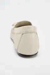 Zapato plano tipo mocasín color crema, confeccionado en cuero, con detalle de lazo con pasador y fruncido en la parte delantera. Pespunte en el corte y acabado en punta redonda.