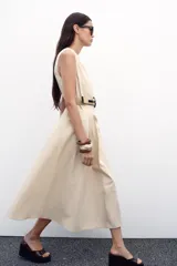 Vestido midi color beige, de popelín, con cuello redondo y sin mangas. Presenta cintura entallada con pinzas, cinturón y falda evasé.