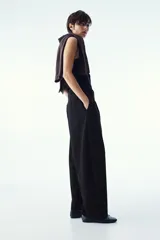 Pantalón negro de corte recto y holgado, con textura de sarga y tiro alto. Tiene detalle en V en la parte posterior de la pretina, cierre con botón y corchete ocultos, piernas anchas con pinzas adelante, bolsillos laterales discretos y bolsillos de atrás de parche.