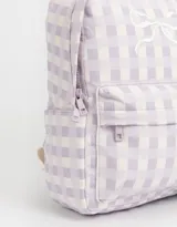 Mochila de tela con estampado a cuadros lila y blanco, con bolsillo frontal con cierre y moño bordado. Tiene correas regulables, manija superior y doble cierre superior.