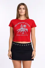 Remera roja de manga corta y corte cropped (baby tee) con cuello redondo. Estampado frontal en azul claro que incluye el texto "DELOALTO CLUB", una figura de sirena montando a caballo sobre una palmera y el texto "Deloutto mvdeo est. 2015".