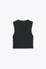 Top corto negro de punto canalé, con cuello redondo y sisa. Presenta detalle de botones metálicos irregulares en los laterales.