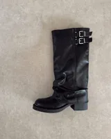 Bota alta de cuero vegano color marrón oscuro, con caña arrugada y detalle de dos hebillas ajustables en la parte superior. Posee suela y taco bajo de goma negra.