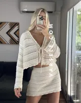 Conjunto de dos piezas tejido al crochet color crudo. Incluye una mini falda de tiro alto y un cardigan corto con mangas largas y lazos para atar en el frente.