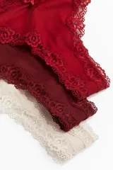 Pack de tres bombachas tipo tanga de algodón acanalado en color crudo, rojo y borgoña, con ribetes de encaje y moño decorativo.
