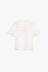 Top de tejido satinado color blanco, con cuello redondo, manga corta y detalle de puntilla a tono. Cierre frontal con lazadas.