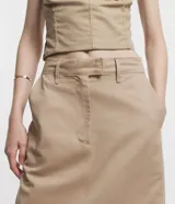 Pollera larga color beige, de tiro alto, corte recto y confeccionada en sarga ligera. Presenta trabillas en la cintura, cierre con botón y cremallera, y abertura en la parte posterior.