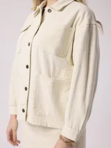 Campera de jean color beige, de corte recto, con cuello clásico, cierre frontal con botones metálicos, bolsillos de parche en el pecho y mangas largas con puños abotonados.