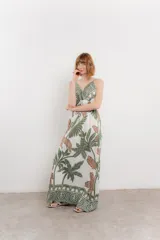 Vestido largo con estampado de hojas en tonos verdes y marrones sobre fondo blanco. Tiene breteles finos, escote en V y detalle de lazo en el frente. La falda es amplia y con vuelo.