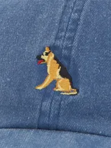 Gorra de algodón orgánico azul con bordado de un pastor alemán en el frente. Tiene visera curva, seis paneles, forro interior de algodón orgánico y ajuste trasero con pasador metálico.