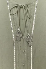 Blusa verde oliva de algodón con cuello redondo y escote en pico con cordones y borlas. Tiene manga larga con bordados florales en los hombros, jaretas y entredoses delanteros. Cierre frontal con botones.