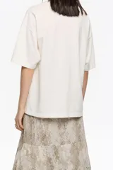 Camiseta blanca de algodón orgánico, corte oversize, con escote redondo y mangas cortas anchas. Presenta un logo grande en relieve en el frente.