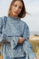 Camisa celeste con estampado floral blanco, cuello alto con volados, mangas largas abullonadas con puños elásticos y corte a la cintura con volados.