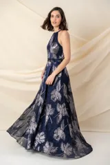 Vestido largo de fiesta en gasa plisada color azul con estampado plateado de hojas. Tiene falda amplia, cruzada y abierta, con bolsillos laterales. El escote es halter y tiene un cinto fino a la altura de la cintura.