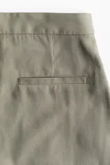 Pantalón color verde oliva de tiro alto con cierre, botón y corchetes ocultos, bolsillos al bies y bolsillos atrás decorativos. Piernas amplias con pliegue adelante y atrás.