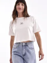 Remera corta de manga corta, color blanco, con cuello redondo y detalles de costura en los hombros y mangas. Presenta un pequeño texto estampado en el pecho.