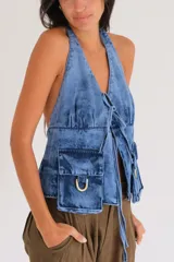 Chaleco tipo top de denim con efecto ahumado (lavado ácido/tie-dye en tonos azules y morados). Presenta escote marcado, bolsillos tipo cargo frontales con argollas metálicas en color bronce y cordones ajustables en el frente.