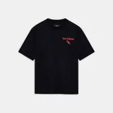 Camiseta negra de corte regular con cuello redondo y manga corta. Presenta un estampado frontal en color rojo/rosado con el texto "Amiri Hollywood" en tipografía gótica y una estrella estilizada con las letras "M" y "A" debajo.