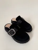 Zueco tipo mule negro, confeccionado en cuero vacuno, con puntera cuadrada y suela track gruesa. Presenta una faja ancha con hebilla metálica plateada sobre el empeine.