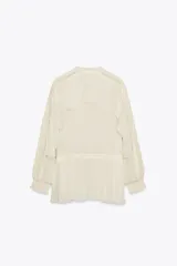 Blusa color crudo confeccionada en viscosa, con cuello redondo, escote en pico, mangas largas con puños elásticos, volantes frontales y detalles de encaje.