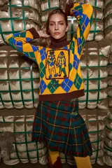Sweater tejido de algodón color amarillo con cuello marrón y diseño de rombos azules y verdes. Presenta un bordado frontal de dos figuras humanas. De corte holgado con cuello a la base y puños.