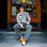 Camisa de hombre de corte boxy fit, con estampado de rayas verticales en tonos grises y blancos, confeccionada en gabardina de algodón. Presenta cuello clásico, mangas cortas y bordado con el logo de la marca en la espalda.