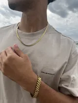 Conjunto de joyería de color dorado, compuesto por una cadena de eslabones tipo cubana o espiga y una pulsera a juego con eslabones anchos y planos. El conjunto se luce sobre una camiseta beige de manga corta con texto estampado en negro.