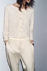 Chaqueta color crema de tejido de punto con lentejuelas, cuello barco, manga larga y cierre frontal con botones.