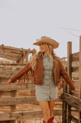 Chaqueta de cuero color marrón con corte entallado, cierre frontal asimétrico con cremallera, cuello con solapas de muesca y flecos decorativos en las mangas.