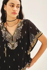 Blusa de gasa traslúcida negra con escote en V y mangas amplias de largo irregular, bordada con canutillos, lentejuelas y mostacillas doradas. Incluye gancho oculto en el escote y viso al tono con breteles regulables y puntilla en el escote.