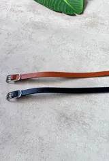 Cinto fino de cuero sintético, disponible en color negro y marrón. Presenta una hebilla metálica plateada de forma rectangular.