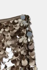 Falda corta de corte recto, completamente cubierta de lentejuelas redondas en tonos plateados y marrones/bronce. Posee forro interior y cierre lateral oculto.