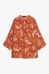 Camisa arrugada de ramio color naranja con estampado floral en tonos rosados y blancos. Tiene cuello redondo con escote pico ajustable con lazadas, mangas 3/4 y bajo asimétrico con aberturas laterales.