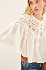 Camisa blanca de algodón con cuello redondo, corte amplio y mangas cortas abullonadas. Presenta detalles de calado en la parte superior delantera y trasera. Cierre frontal con botones.