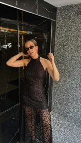 Vestido largo de encaje negro con cuello halter y espalda descubierta. Tiene un corte a la altura de la rodilla y un ruedo largo transparente.
