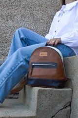 Mochila de cuero color tostado, con base y espalda en negro y tapa superior en blanco. Tiene un bolsillo frontal con cierre y un pequeño aplique de cuero con el logo de la marca.